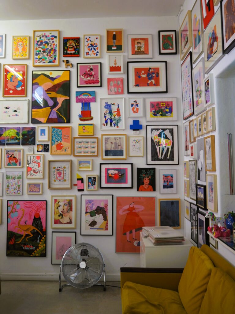 Colourful gallery wall filled with framed art above a yellow sofa and a metal floor fan, reminiscent of the vibrant creativity found at popular attractions in Porto.