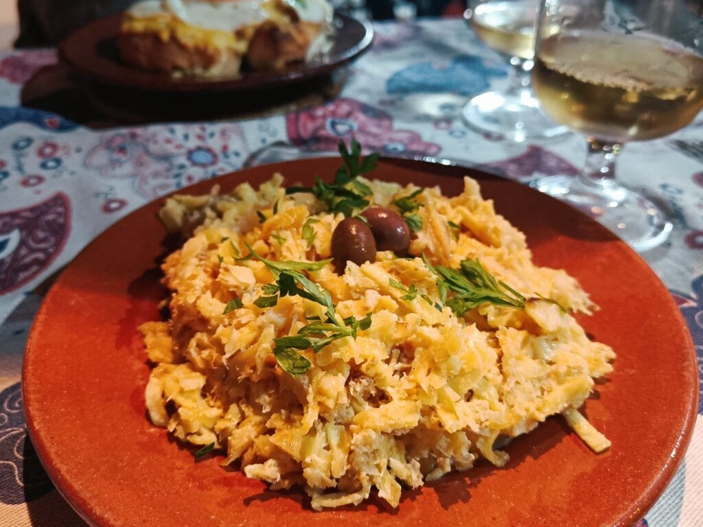 A plate of bacalhau à Brás garnished with olives and parsley, a classic dish among the best food Porto has to offer, with wine glasses in the background.