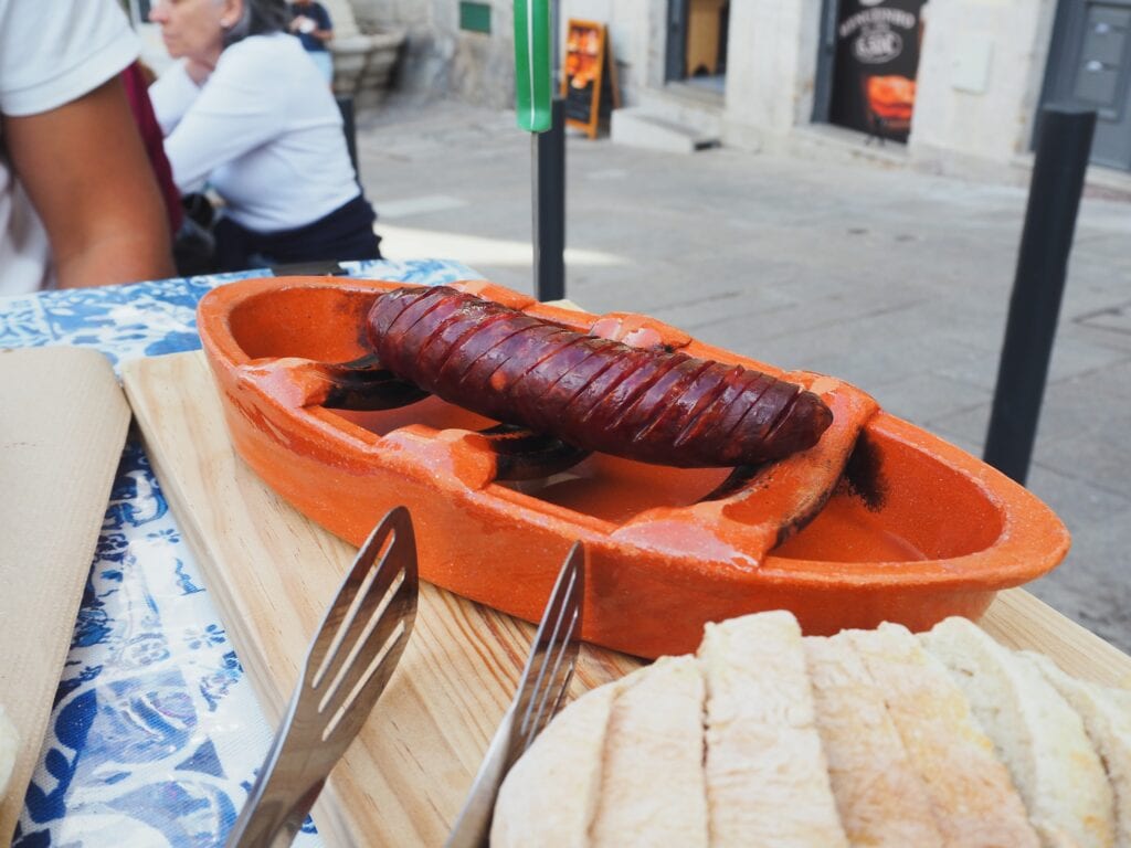 A grilled sausage on a ceramic dish with sliced bread and cutlery on an outdoor table, showcasing the best food Porto has to offer.