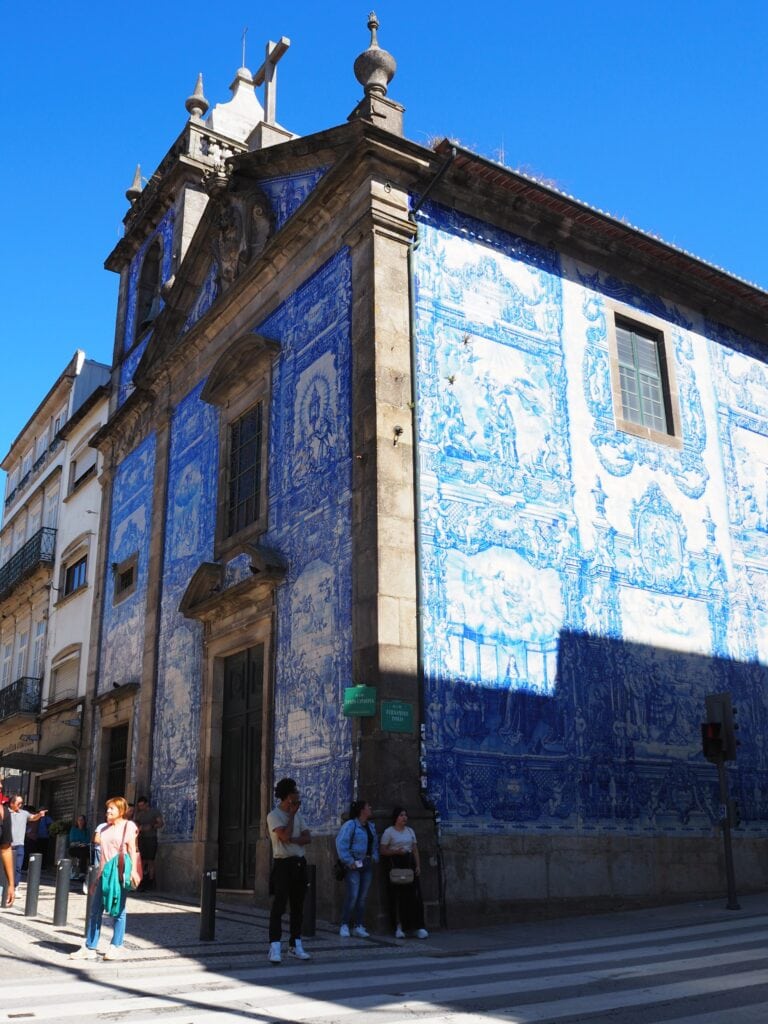 A church with blue and white tile murals on its exterior, people standing and walking nearby under a clear sky—one of the must-see Porto attractions for those exploring free things to do in Porto.