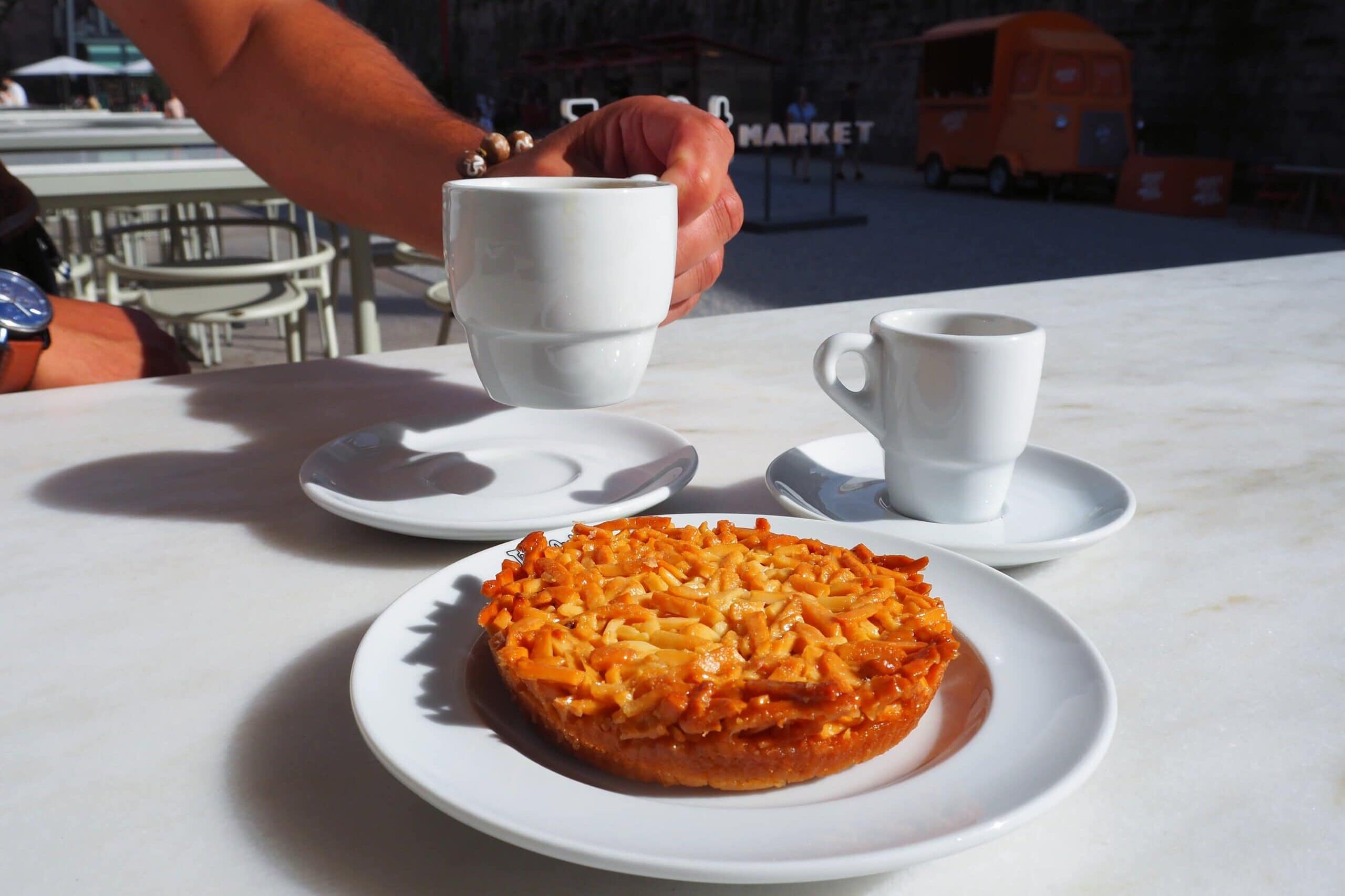 A hand lifts a coffee cup beside a small tart and another cup on a white outdoor table, inviting you to savour the best food Porto has to offer.