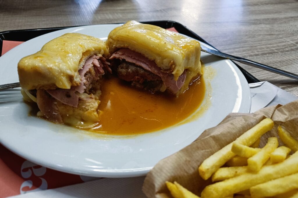 A sandwich topped with melted cheese and sauce, cut in half on a plate, with chips on the side—experience the best food Porto has to offer.