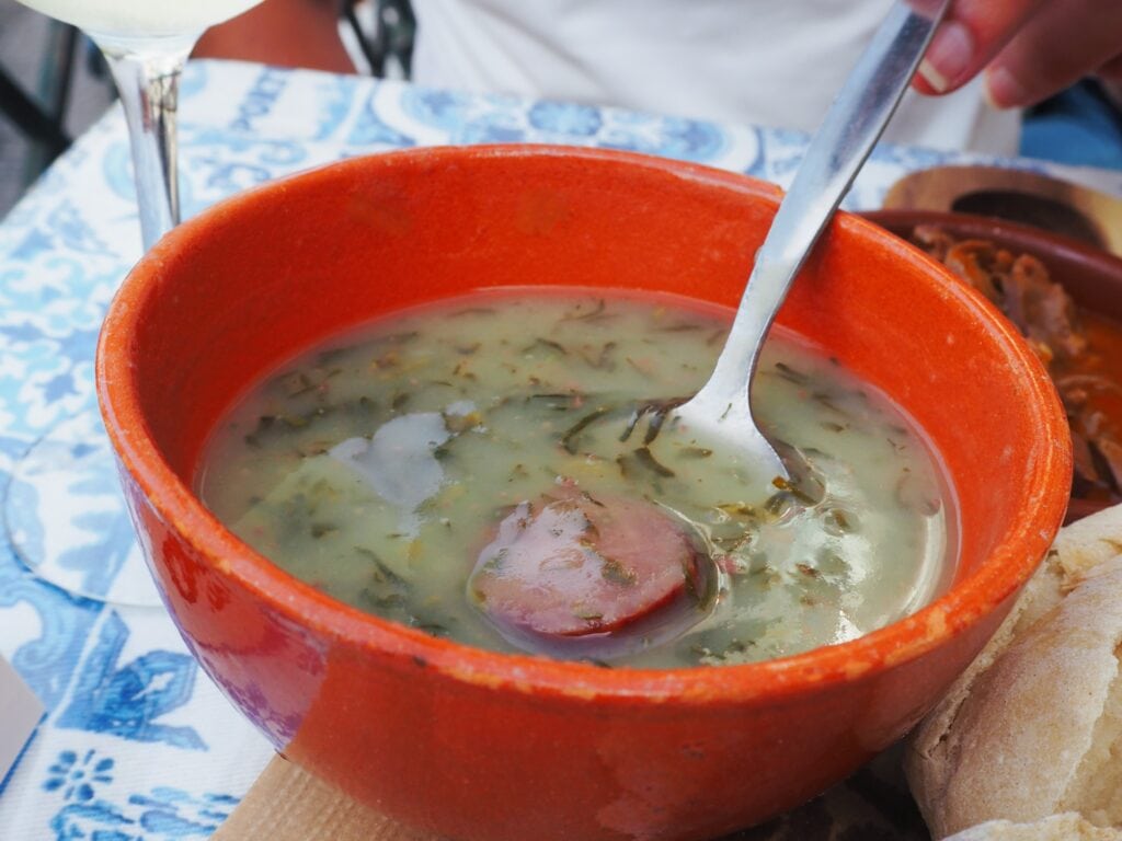 A bowl of green herb soup with sausage slices, served in an orange bowl with a spoon—an example of the best food Porto has to offer.