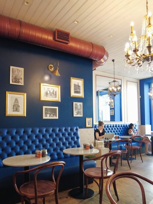 Cosy café with blue tufted seating, chandeliers, framed art on the wall, and people sitting at tables—renowned for serving the best pastéis de nata in Porto.
