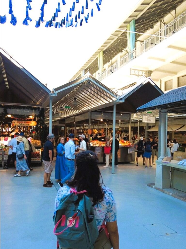 A woman with a rucksack looks at people shopping at a busy indoor market with high ceilings and bright decor—one of the Best Free Things to do in Porto.