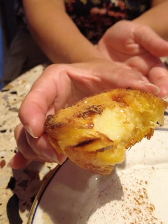 A hand holding a half-eaten, custard-filled pastry over a plate on a speckled worktop.