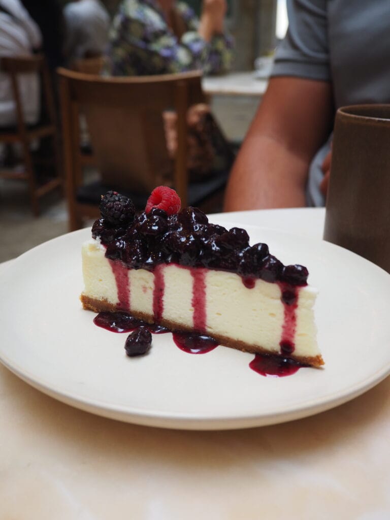 A slice of cheesecake topped with berries and berry sauce on a white plate, perfect for those searching where to eat in Porto, with a blurred background.