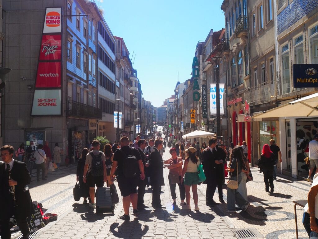 Busy street scene with people walking and gathering, flanked by shops and restaurants on a sunny day—a perfect glimpse into what to do in Porto whilst exploring its popular attractions.