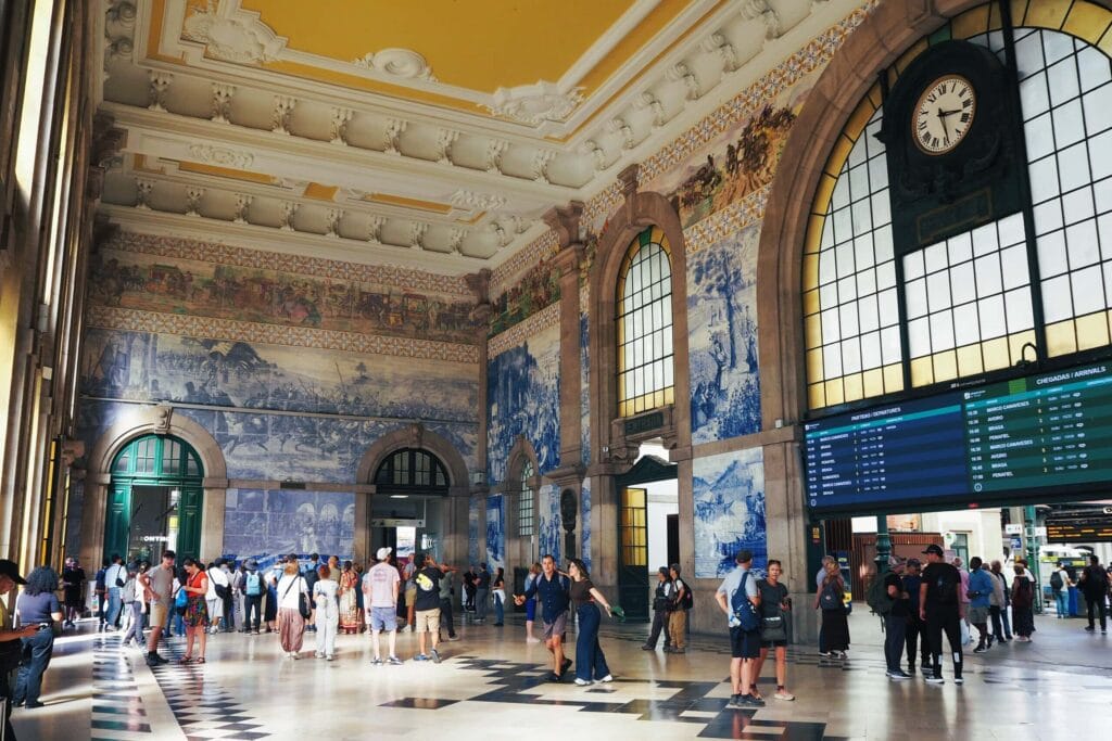 People stand in a grand railway station with large arched windows, blue tiled murals, and a clock above a timetable—one of the Best Free Things to do in Porto.