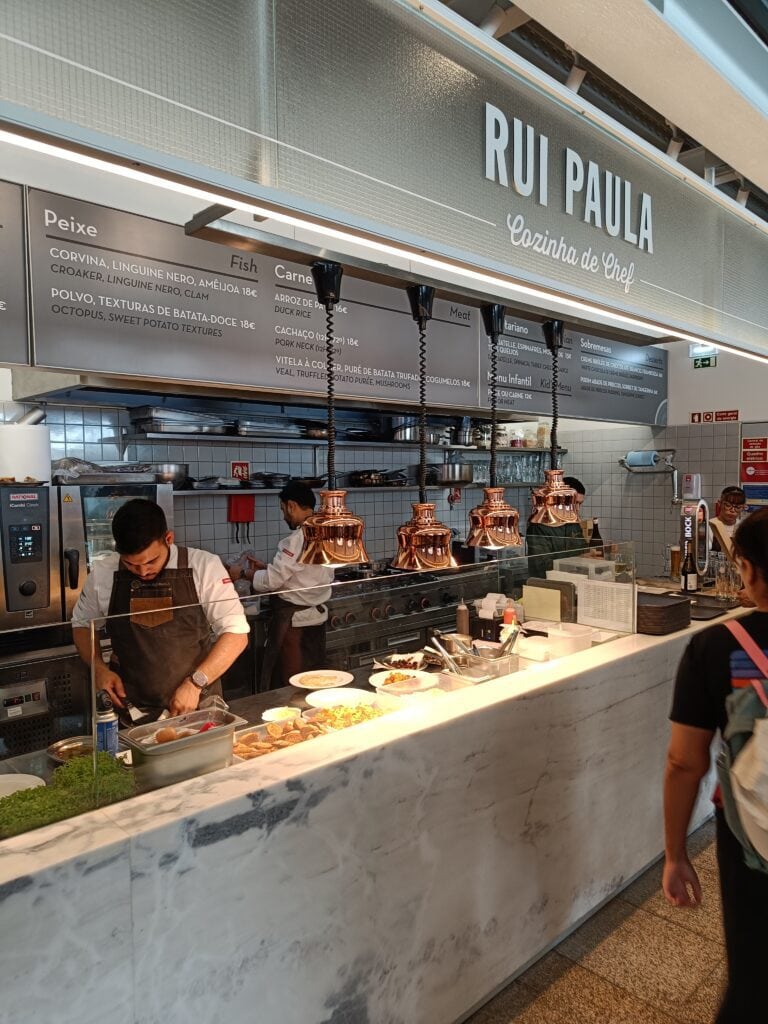 Chef prepares food at a marble counter in Rui Paula restaurant, with menu boards and copper lamps overhead—a must-visit spot if you’re wondering where to eat in Porto.