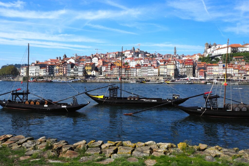 Traditional boats with barrels drift along the river, colourful hillside buildings in the background under a blue sky—one of the popular attractions in Porto.