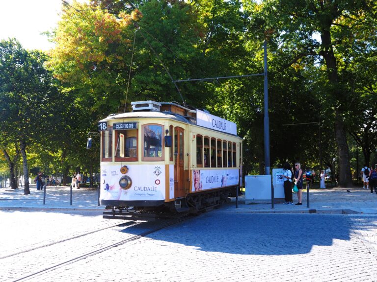 A vintage yellow tram, a classic way of getting around Porto, travels along a cobbled street lined with trees in bright sunlight.