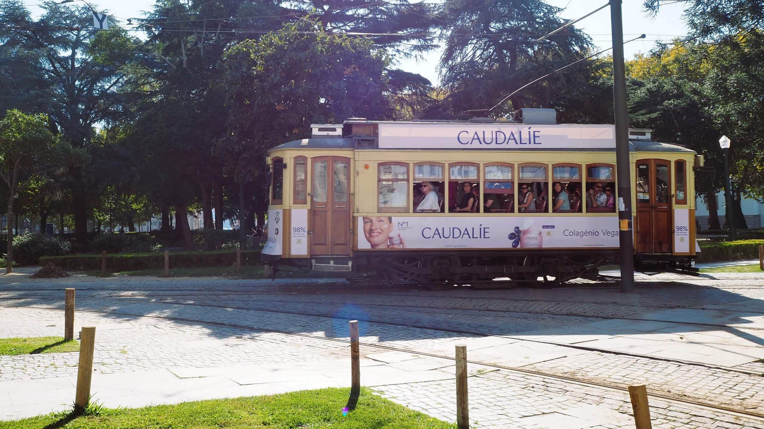 A yellow tram with Caudalie adverts, perfect for getting around Porto, travels past trees and cobbled streets on a sunny day.