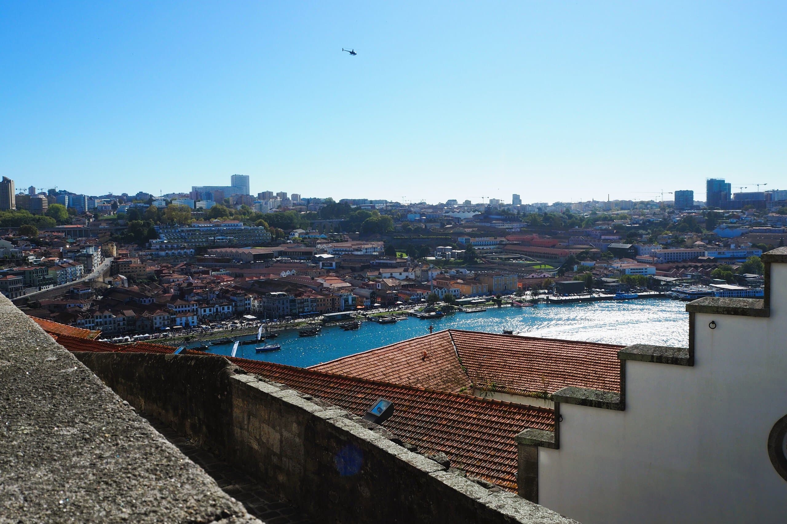 Scenic cityscape with red rooftops by a river under a clear blue sky and a helicopter flying overhead.