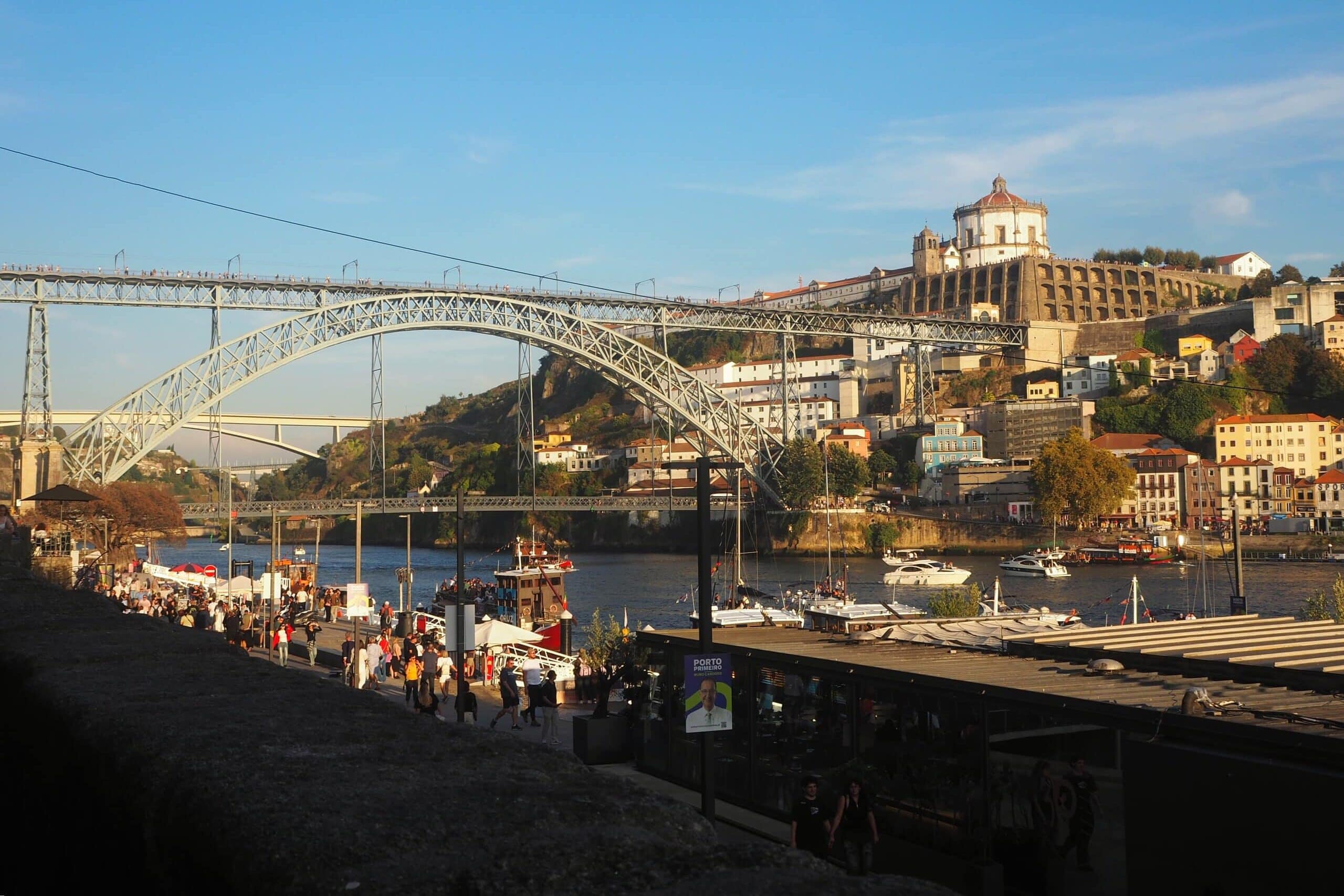 Crowds by the river in Porto, Portugal, enjoy one of the top free things to do in Porto, with stunning views of Dom Luís I Bridge and hillside buildings in the background.