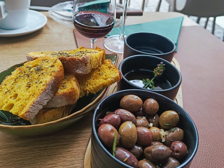 Sliced bread, mixed olives, and olive oil in bowls, with a glass of red wine on a wooden table—enjoying the best food Porto has to offer.