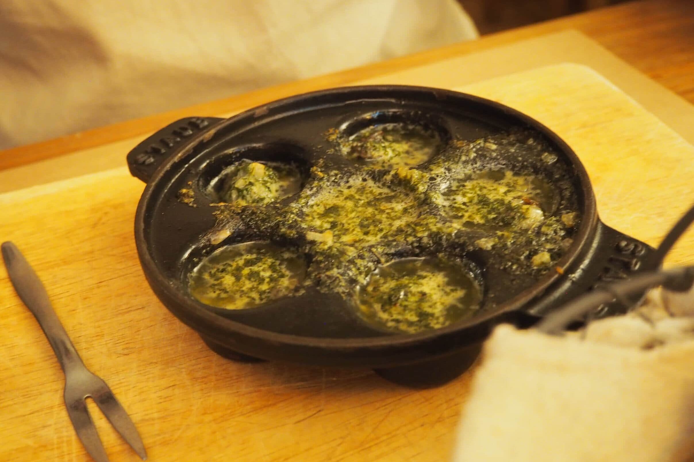 A cast iron escargot dish with cooked snails in garlic butter sits on a wooden board with a small fork nearby—one of the 5x must-try French food experiences for any culinary adventurer.
