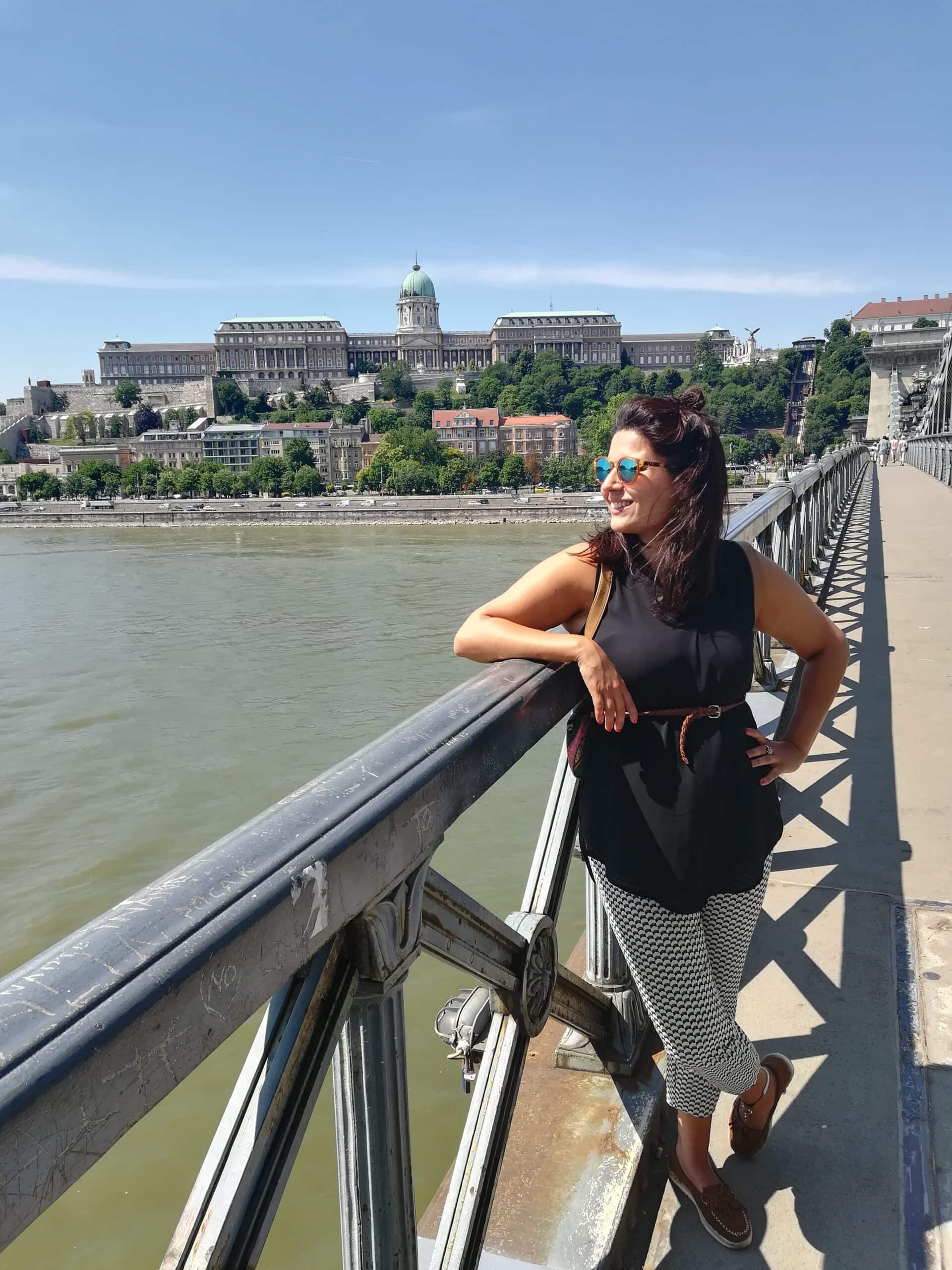 Woman in sunglasses smiling on a bridge over a river, with a large historic building in the background.