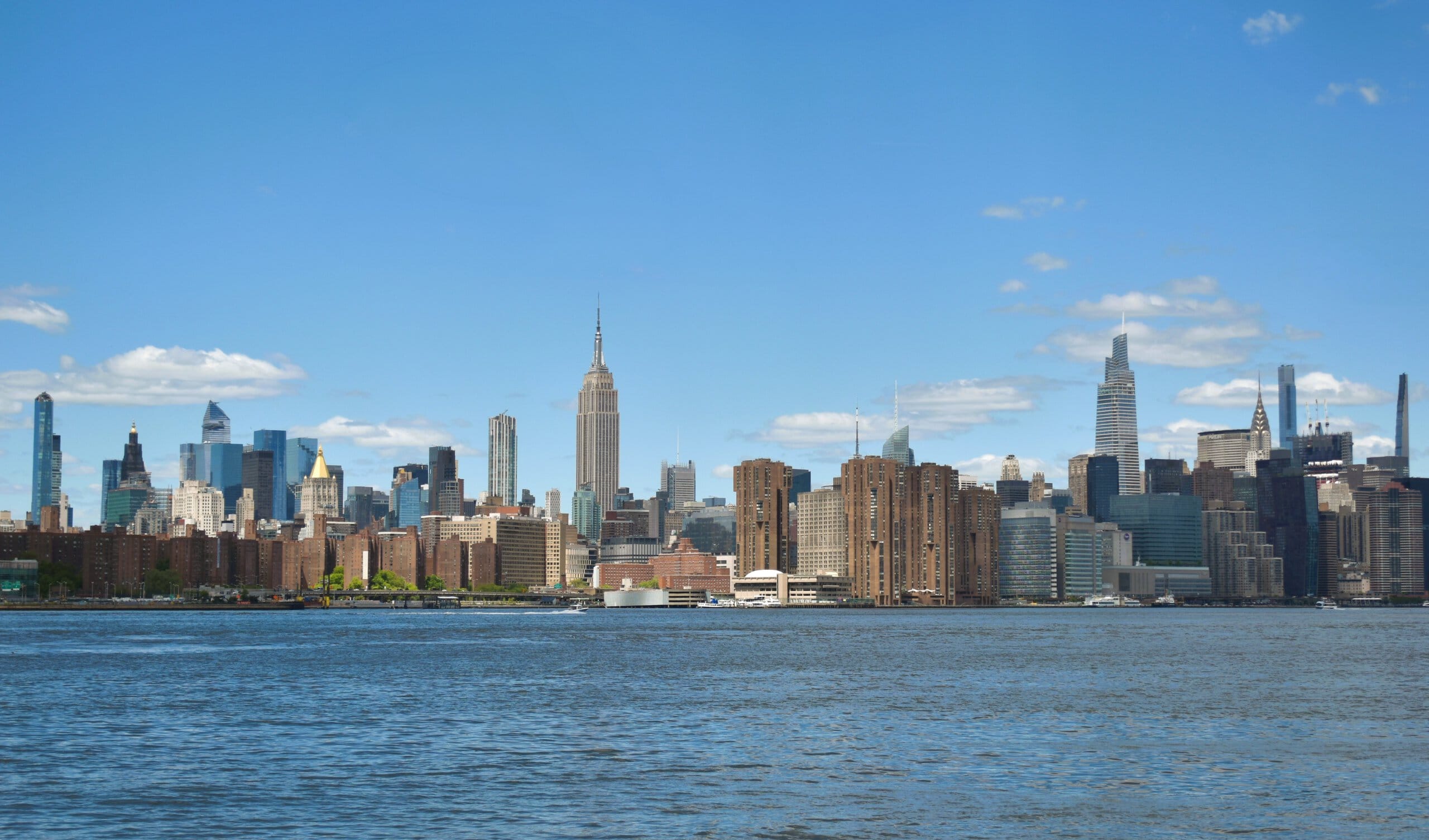 New York City skyline with skyscrapers and blue sky, viewed across the water during the daytime.