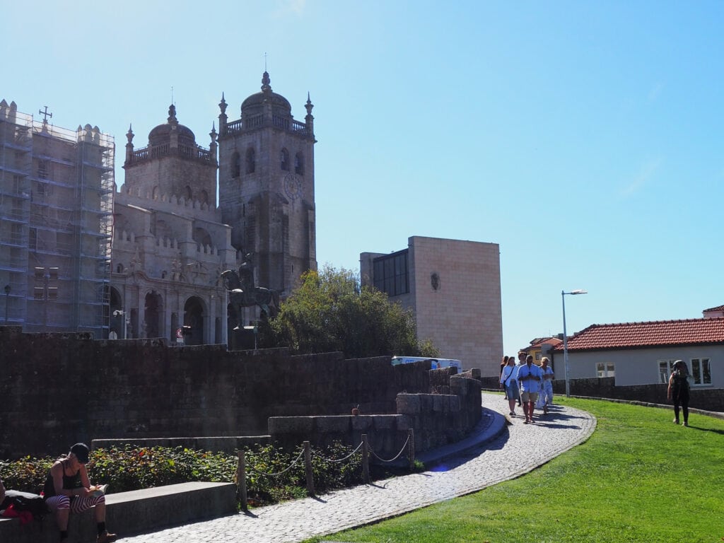 People walk along a cobbled path near a historic stone building with towers on a sunny day, exploring one of the popular attractions in Porto.