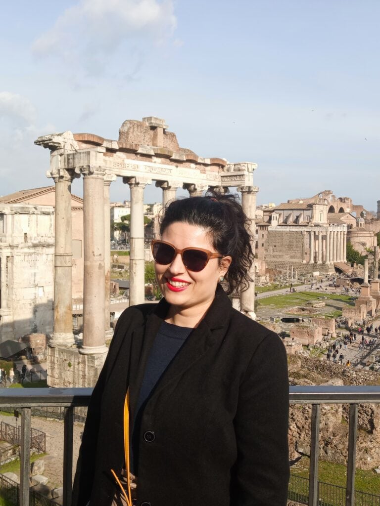 Woman in sunglasses and black coat smiling in front of ancient Roman ruins on a sunny day.