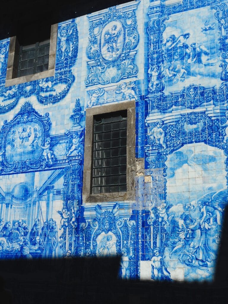 Blue and white tiled mural with intricate designs and portraits surrounds two rectangular windows on a building facade—one of the popular attractions in Porto.