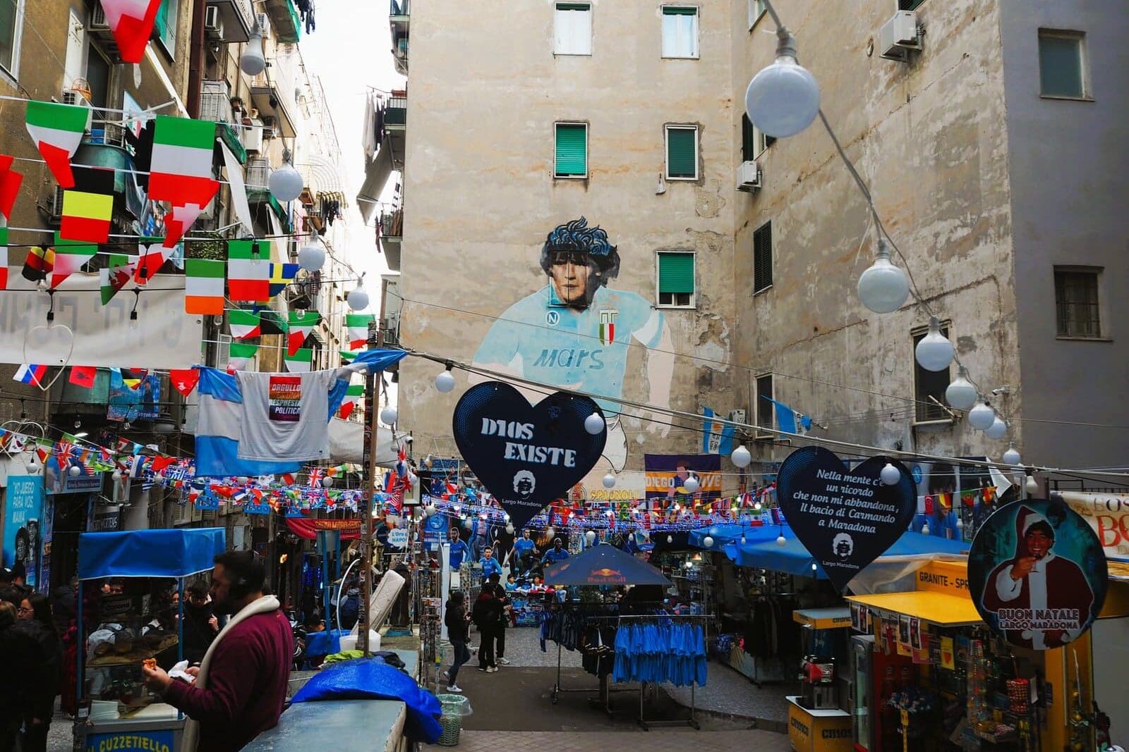 Busy street market decorated with flags, featuring a large mural of Diego Maradona on a building wall.