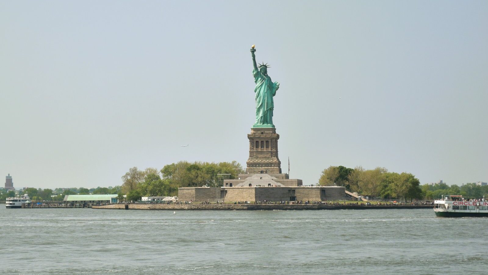 The iconic Statue of Liberty stands majestically on Liberty Island, framed by a clear sky and reflected in the calm water.