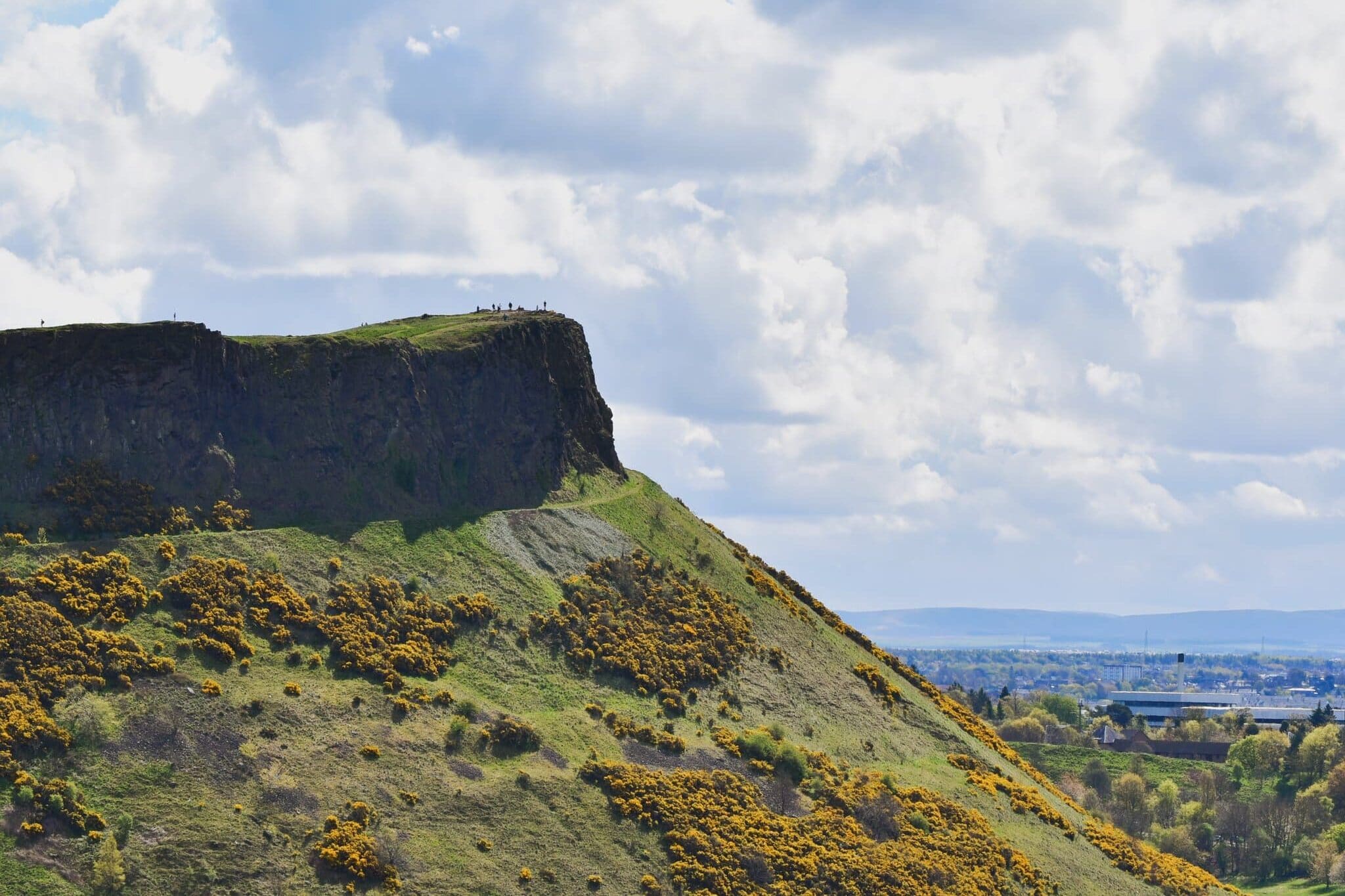 A green hill with a rocky cliff top under a partly cloudy sky, dotted with yellow-flowered shrubs.