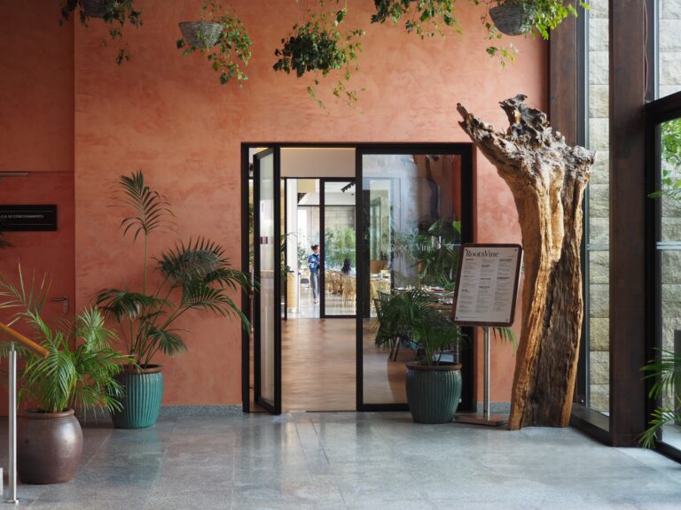 Open glass door in a plant-filled lobby with a tree sculpture and restaurant menu stand near an orange wall—an inviting spot if you’re searching for where to eat in Porto.