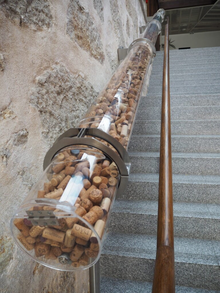 A large clear tube filled with wine corks runs alongside a stone stairway with a wooden handrail, evoking the charm of the World of Wine Porto.
