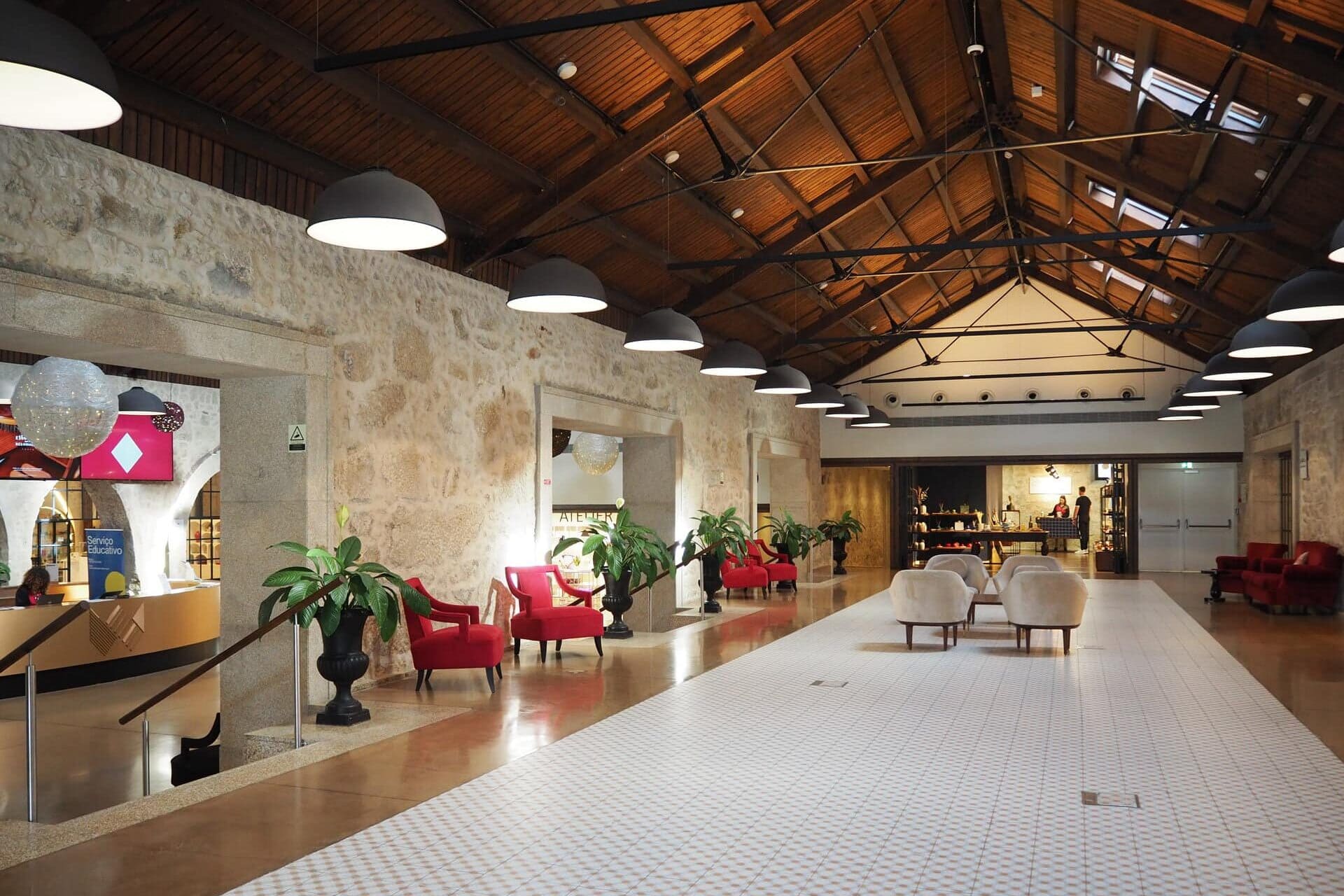 Spacious hotel lobby with stone walls, red chairs, plants, and a high wooden ceiling with hanging lights—perfect for relaxing after exploring WOW Porto and the vibrant World of Wine Porto district nearby.