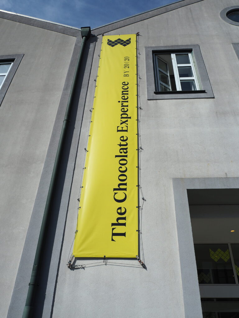 Yellow vertical banner on grey building reads “The Chocolate Experience by XPLXO,” located near WOW Porto. Window open nearby.