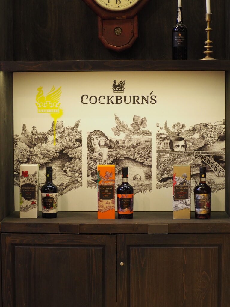 Bottles of Cockburn's port and Tips Porto sit on a wooden shelf, framed by an illustrated backdrop and a vintage clock above.