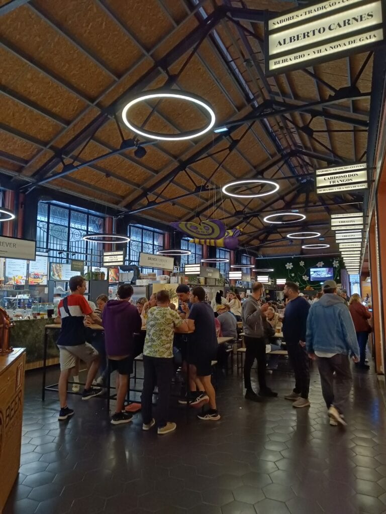 People gather and dine at high tables in a lively indoor market, reminiscent of popular food markets Porto is known for, with food stalls and bright circular lights.
