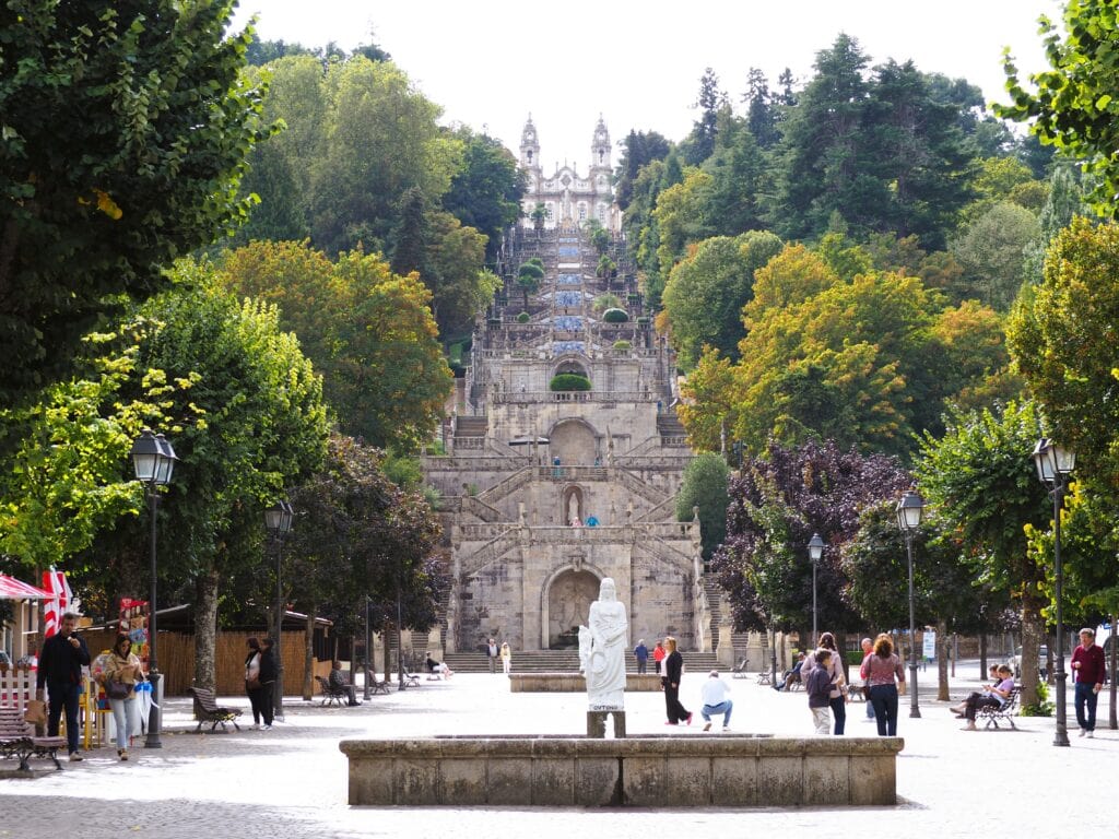 People walk in a square with a large staircase and ornate church surrounded by trees in the background, making it one of the Best Day Trips from Porto.