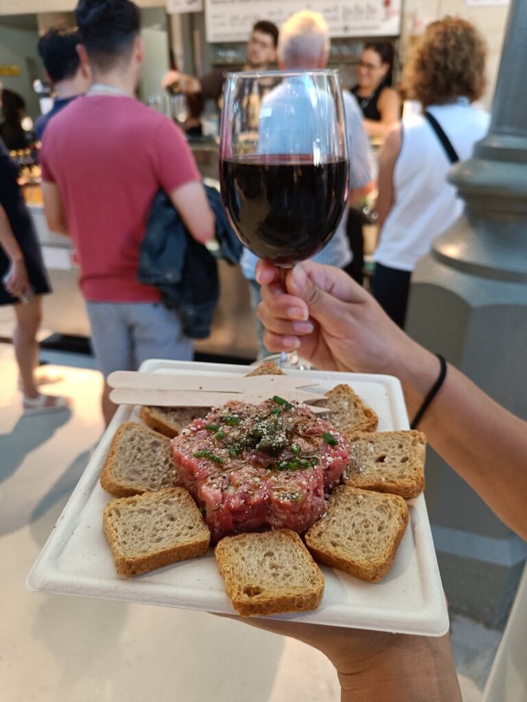 A hand holds a glass of red wine and a plate of steak tartare with toast, surrounded by people at one of Porto's popular food markets indoors.