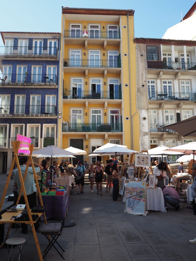 Outdoor art market with stalls and people in front of colourful Porto flat buildings on a sunny day, capturing the lively spirit of local food markets.