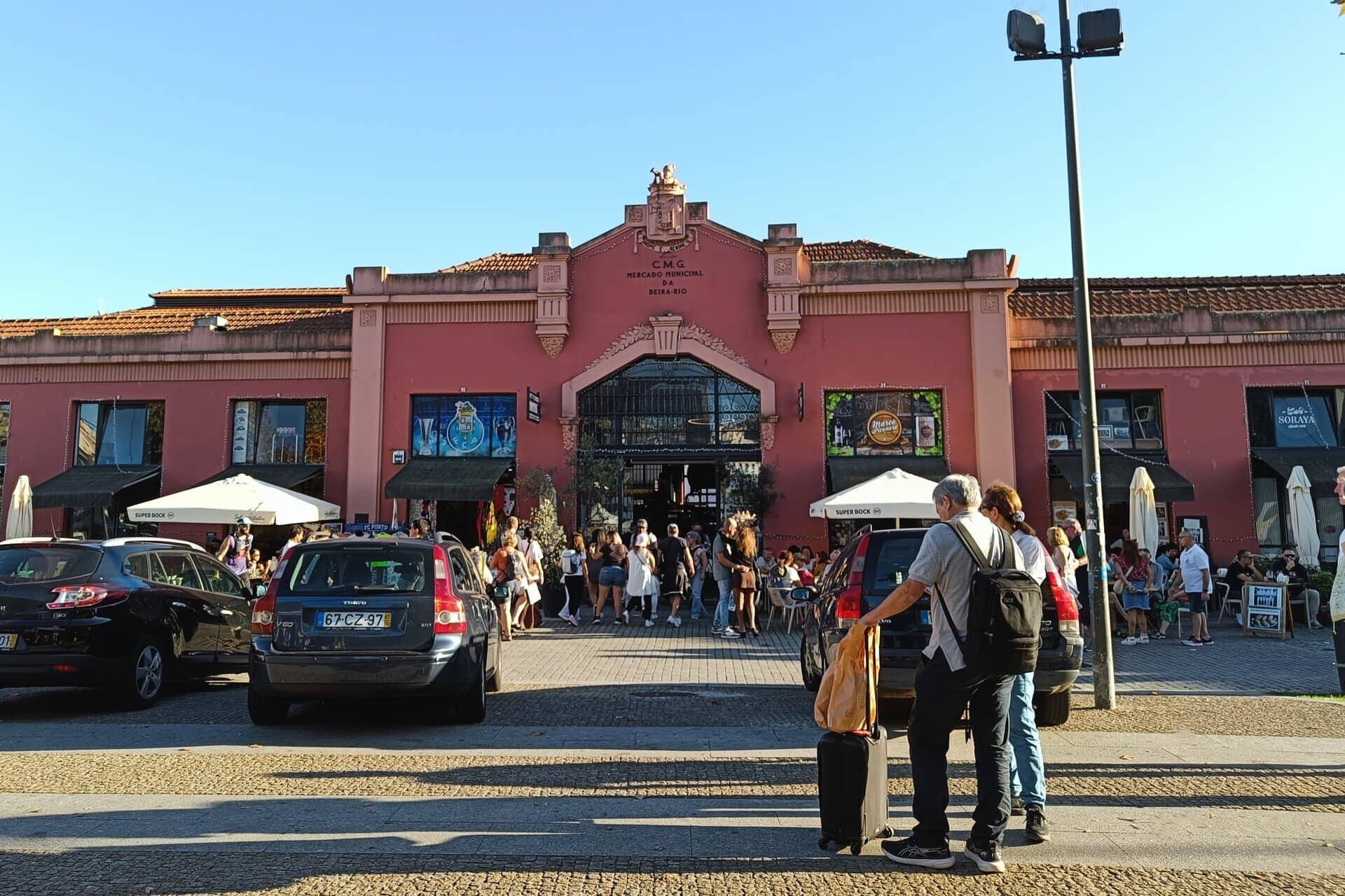 People gather outside a pink building with outdoor seating and parked cars under a clear blue sky, capturing the lively atmosphere often found around Porto markets.