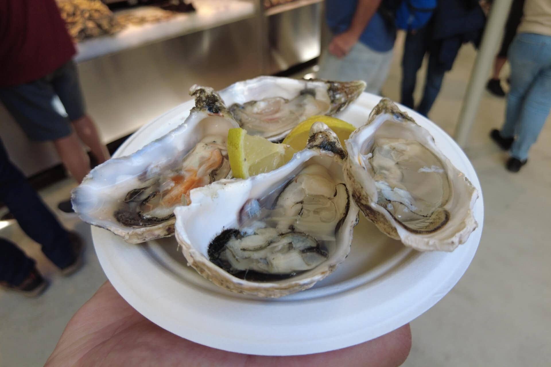 A hand holds a plate of opened oysters with lemon slices; people stand in the background, evoking the vibrant atmosphere found at the best markets in Porto.