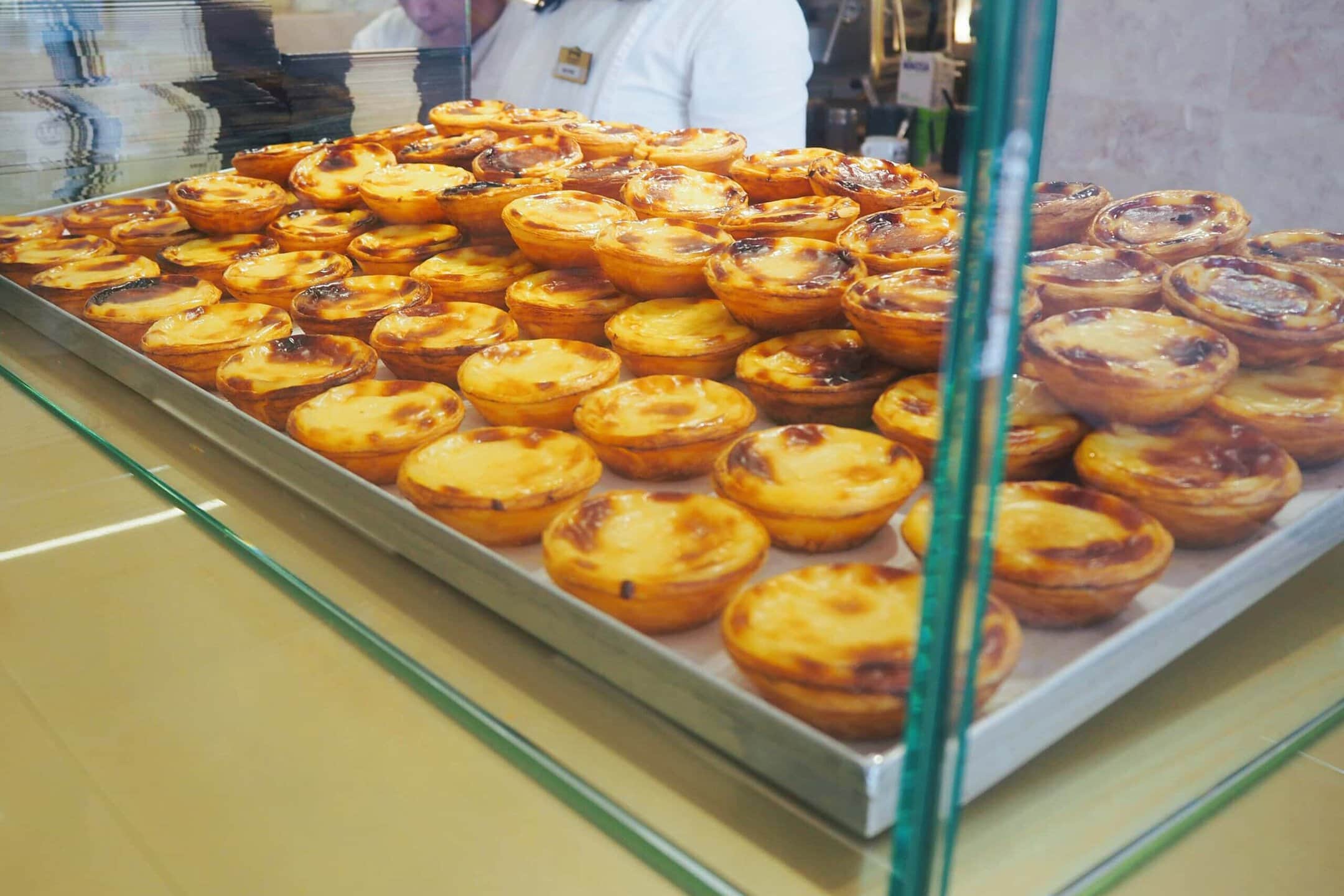 A tray filled with many Portuguese custard tarts, said to be the best pastéis de nata in Porto, on display behind glass at a bakery counter.