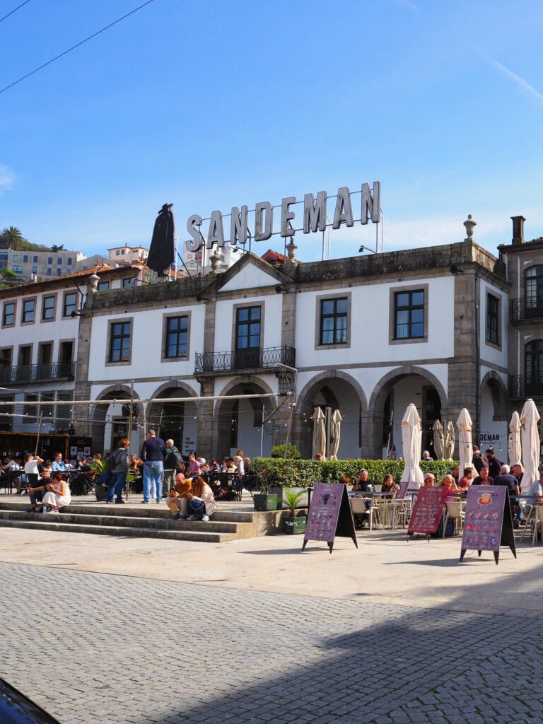 People sit at outdoor tables in front of a historic building with a large "SANDEMAN" sign on the roof, enjoying the atmosphere and exchanging tips about Porto's best sights and experiences.