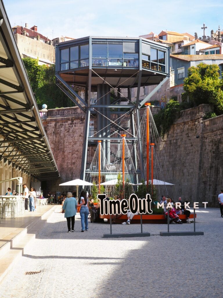 People walking near the Time Out Market sign in Porto, one of the city's popular food markets, with a modern glass lift tower in the background.