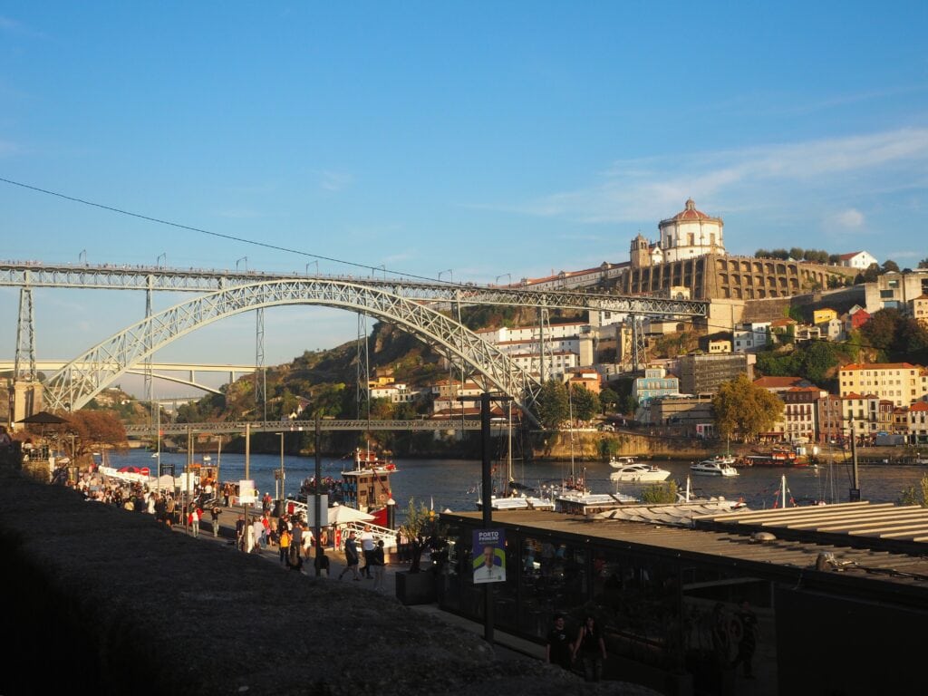 People stroll by the Douro River with the Dom Luís I Bridge and colourful Porto buildings in the background—perfect for photos and discovering local gems with insider tips Porto offers.