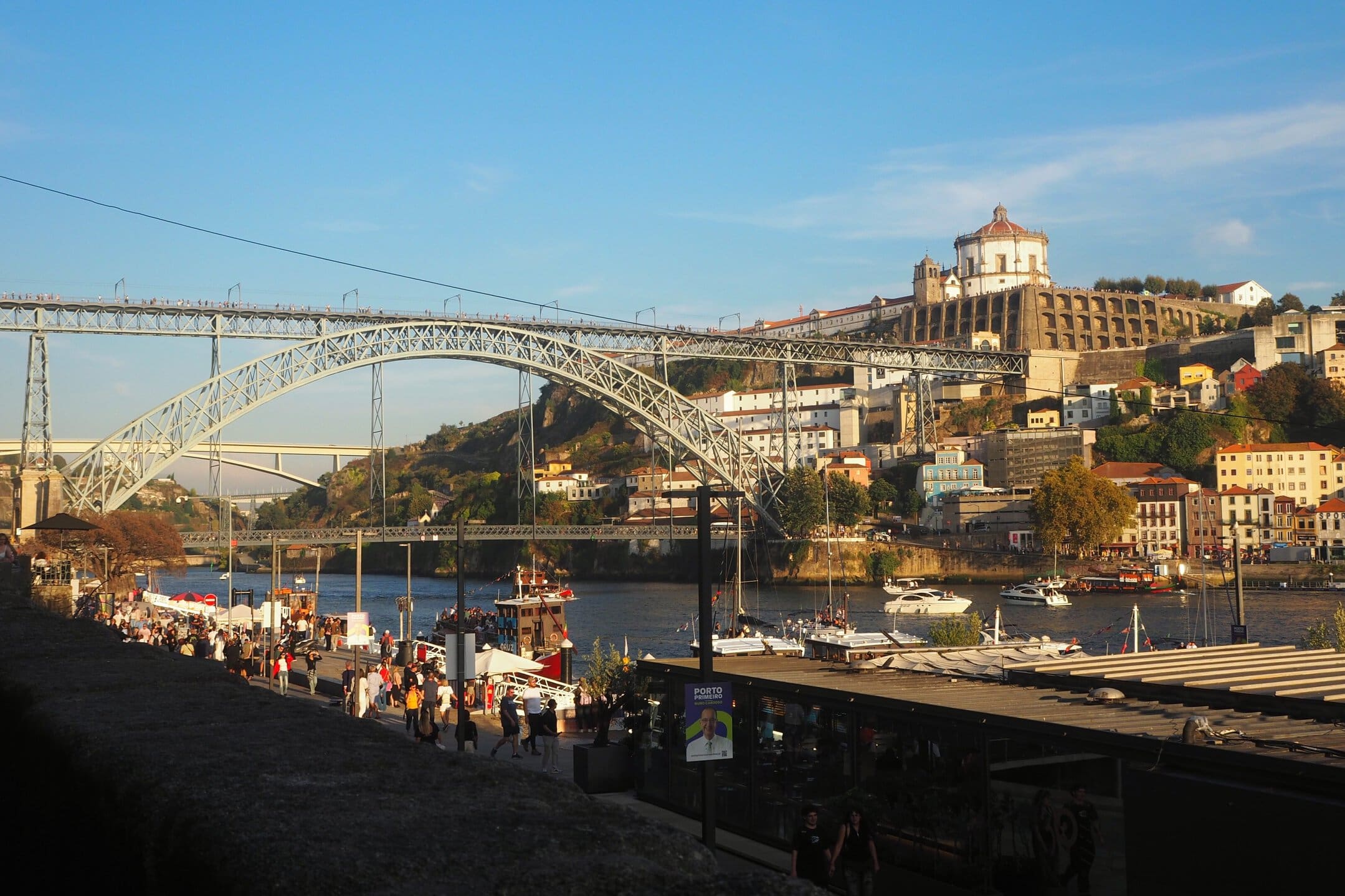 People stroll by the Douro River with the Dom Luís I Bridge and colourful Porto buildings in the background—perfect for photos and discovering local gems with insider tips Porto offers.