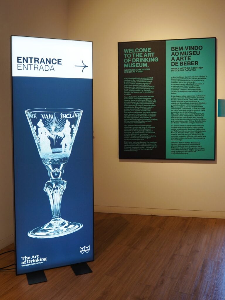 Entrance sign with an ornate glass and a welcome panel at The Art of Drinking Museum on a wooden floor in WOW Porto’s World of Wine district.