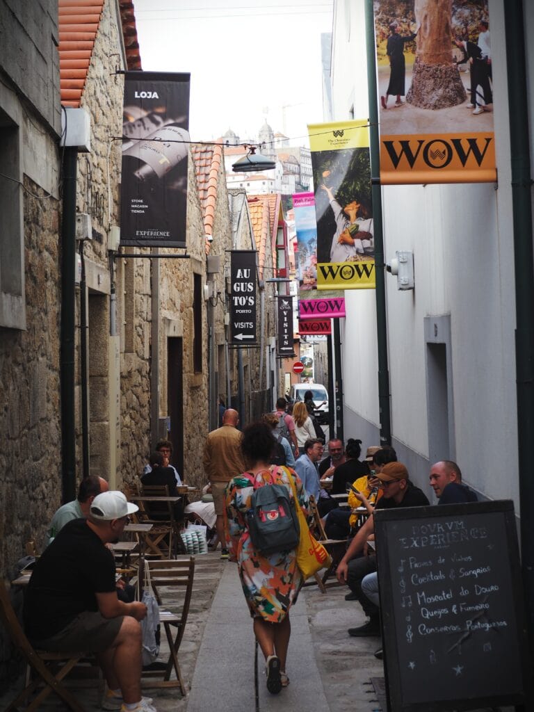 Narrow cobbled alley with people sitting at tables, colourful signs, and a woman walking away—capturing the vibrant charm near WOW Porto in the heart of the World of Wine Porto district.