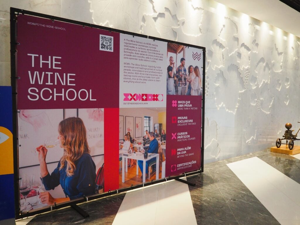 Large sign for "The Wine School" with photos of people enjoying wine classes, displayed in a modern corridor—perfect for those keen to explore the world of wine Porto style.