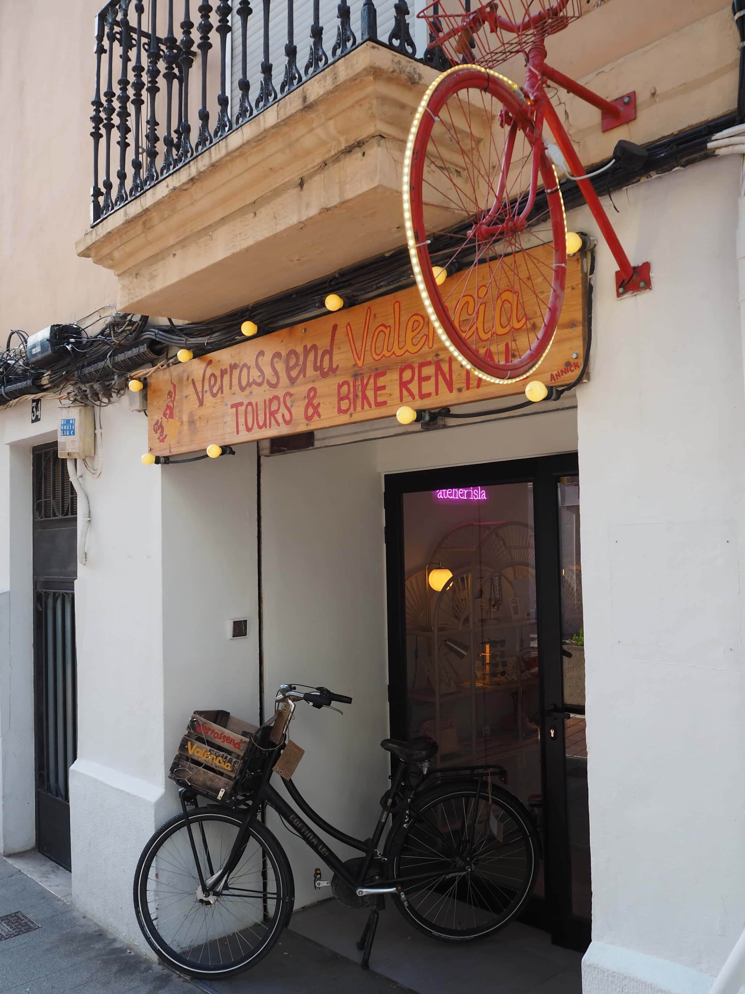 A black bicycle is parked outside a tour and bike hire shop with a red bike mounted above its entrance—a great spot to learn how to get around in Valencia.
