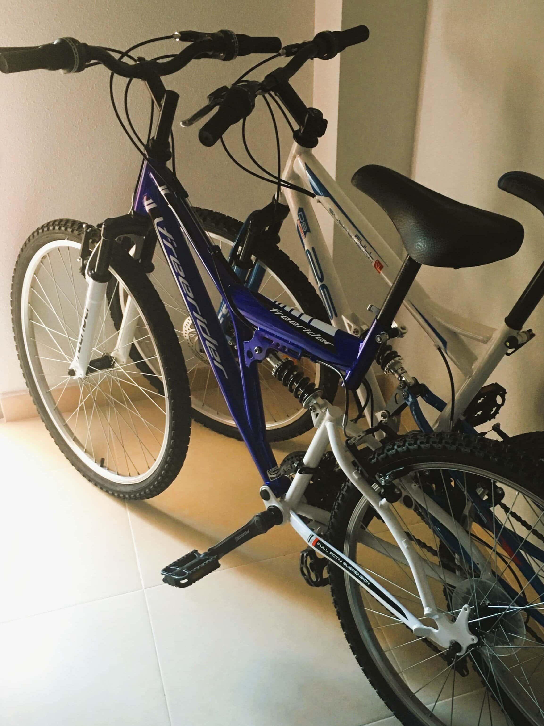 Two bicycles, one blue and one white, are parked indoors side by side on a tiled floor near a wall—showing just one way of how to get around in Valencia.